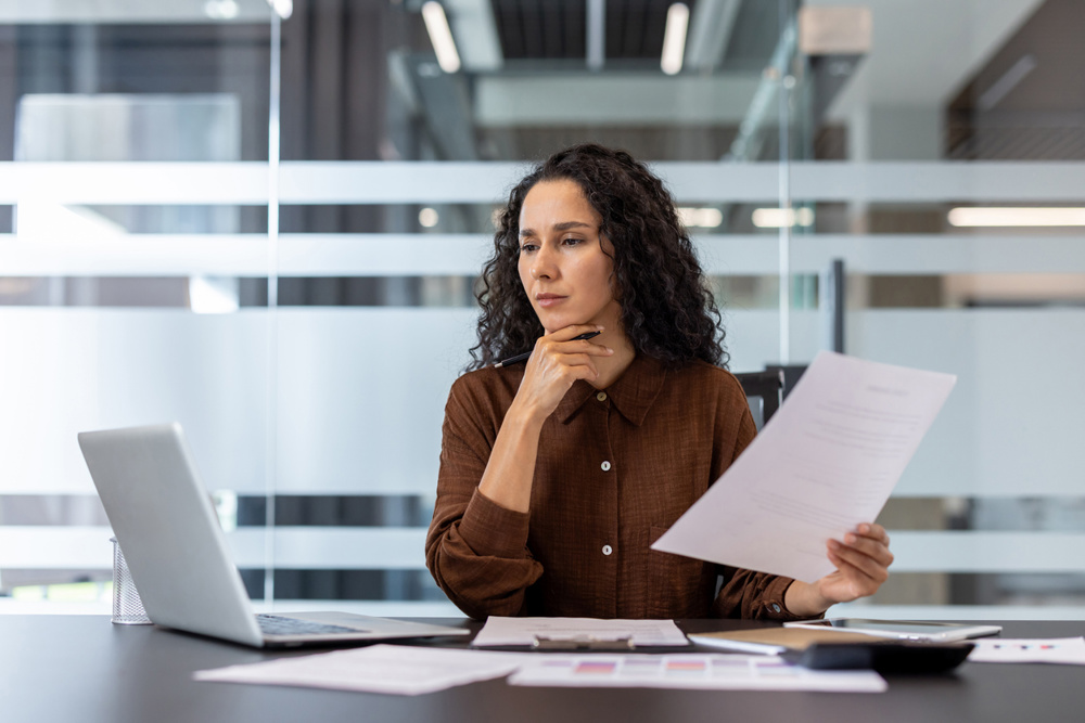 A focused businesswoman is reviewing documents and working on a laptop in a modern office setting. She appears thoughtful